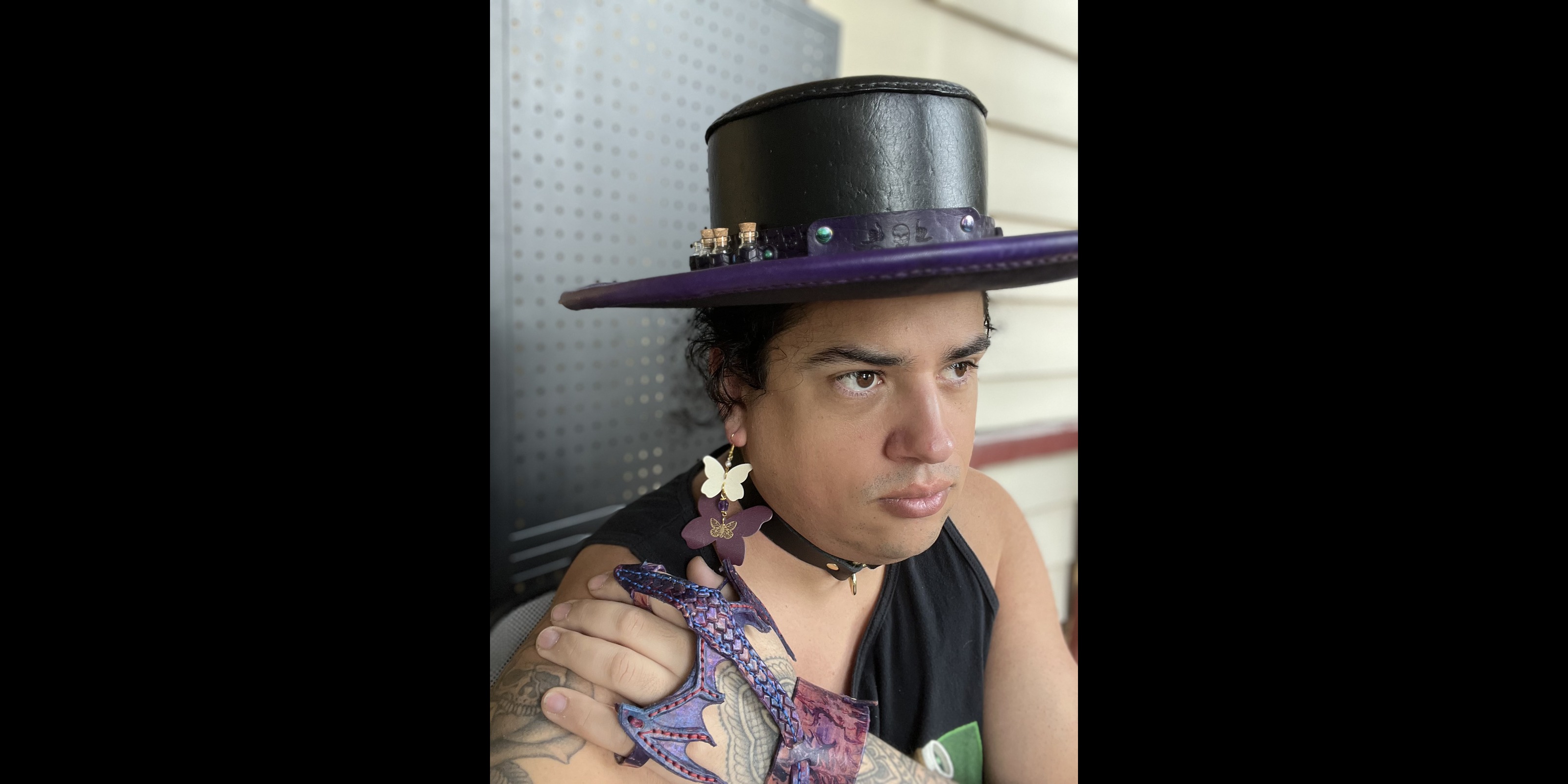 William wearing a handmade leather hat, earrings, choker, and a small leather dragon on their arm